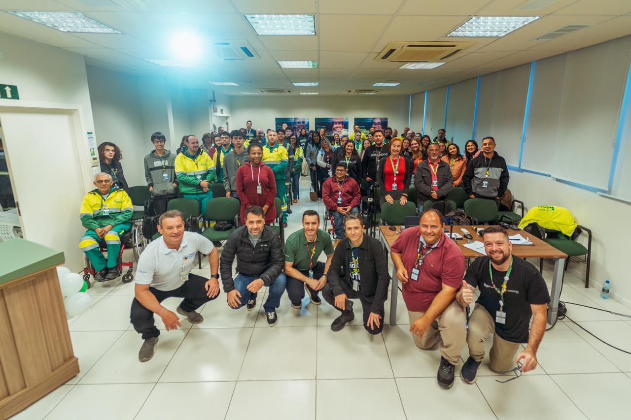 A imagem retrata um grupo de pessoas reunidas em uma sala para uma foto coletiva. Na frente, alguns homens estão agachados, enquanto o restante da audiência está em pé ao fundo, sorrindo para a câmera. Parte dos participantes usa uniformes verdes e amarelos. O ambiente parece ser uma sala de conferências ou treinamentos, com cadeiras e mesas dispostas. 