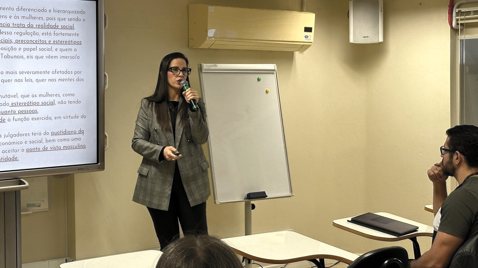 fotografia de uma mulher branca de pé segurando um microfone e falando para o público 