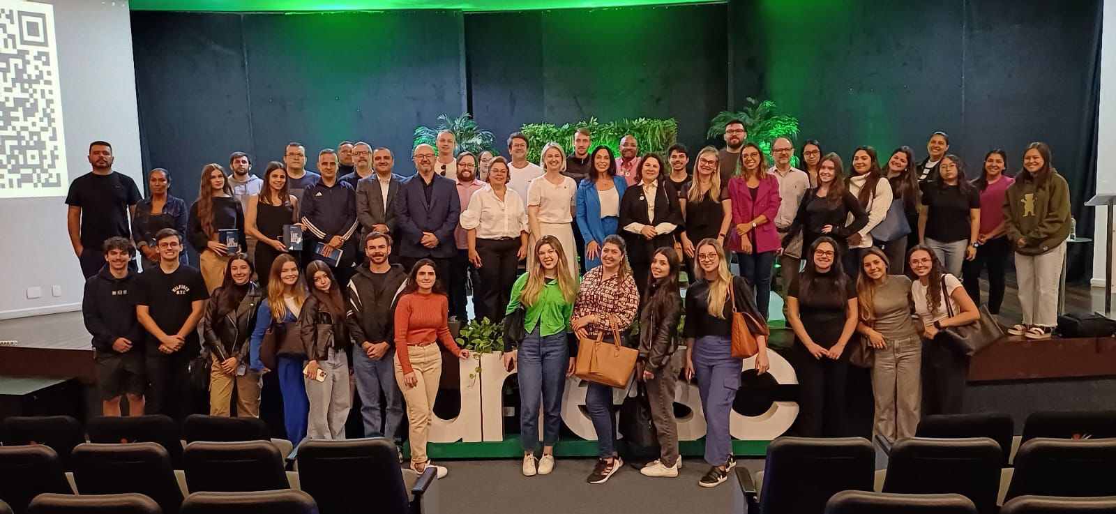 Grupo mostra cerca de 50 pessoas diante de um palco iluminado de verde