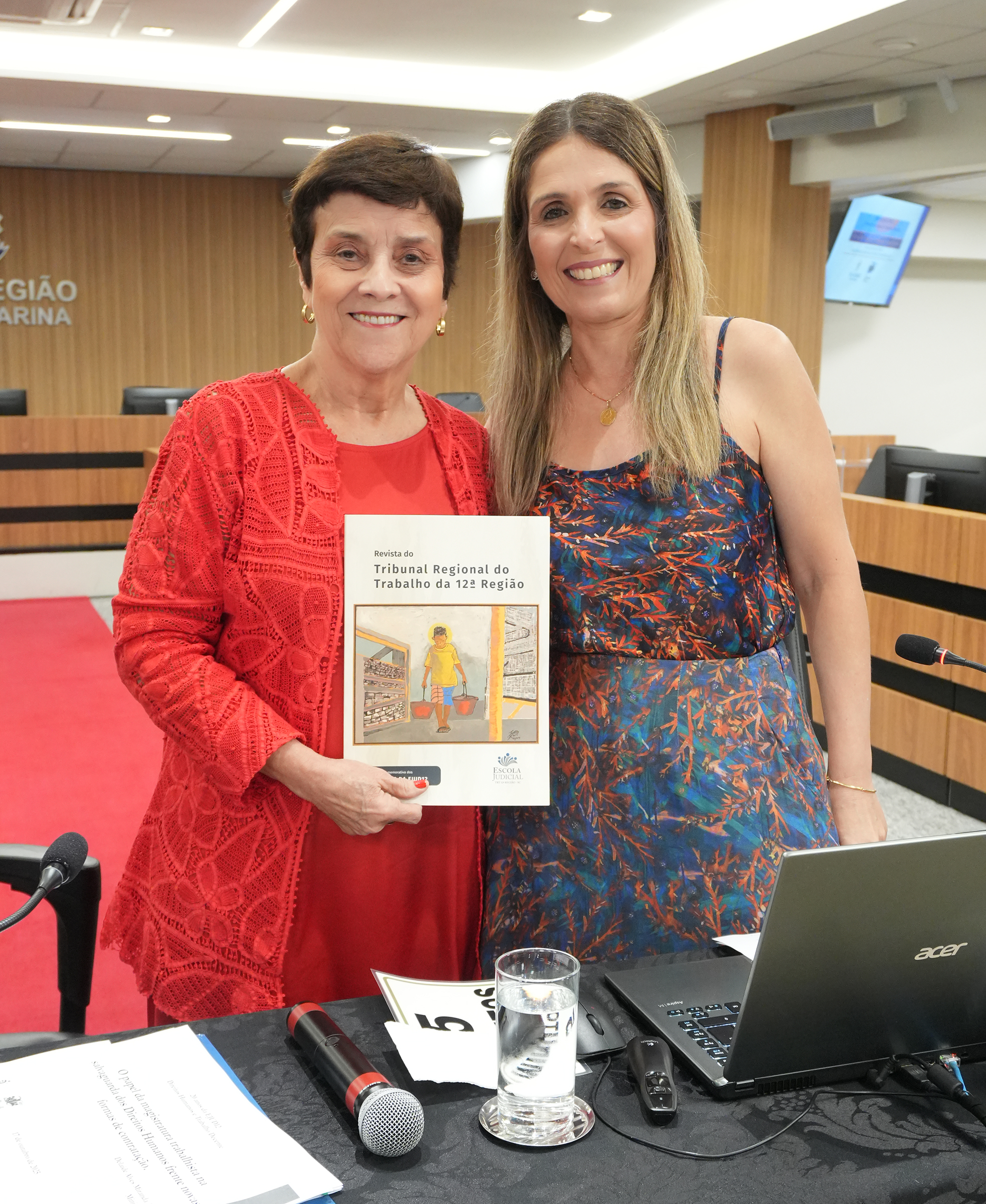 Duas mulheres estão em pé, lado a lado, sorrindo e posando para a foto. Uma delas está segurando uma revista