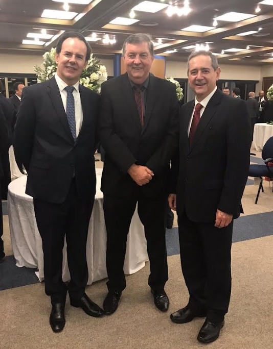 Três homens em pé, de terno e gravata, posam para a foto sorridentes, em um evento