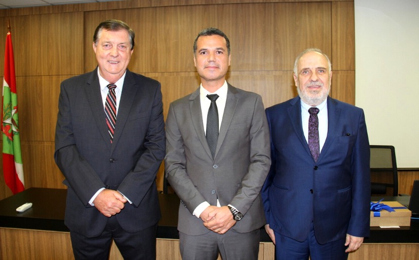 Três homens brancos, em pé, um ao lado do outro, posam para foto. Os três vestem terno e gravata. Ao fundo, uma mesa de trabalho e uma bandeira do Estado de Santa Catarina