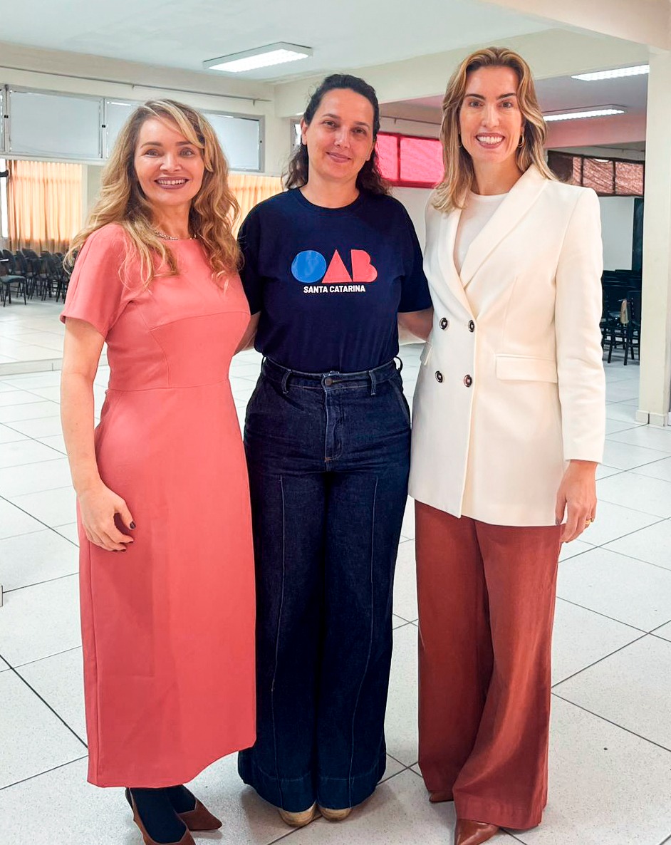 Três mulheres, duas loiras e uma morena, estão em pé sorrindo e posando para a foto