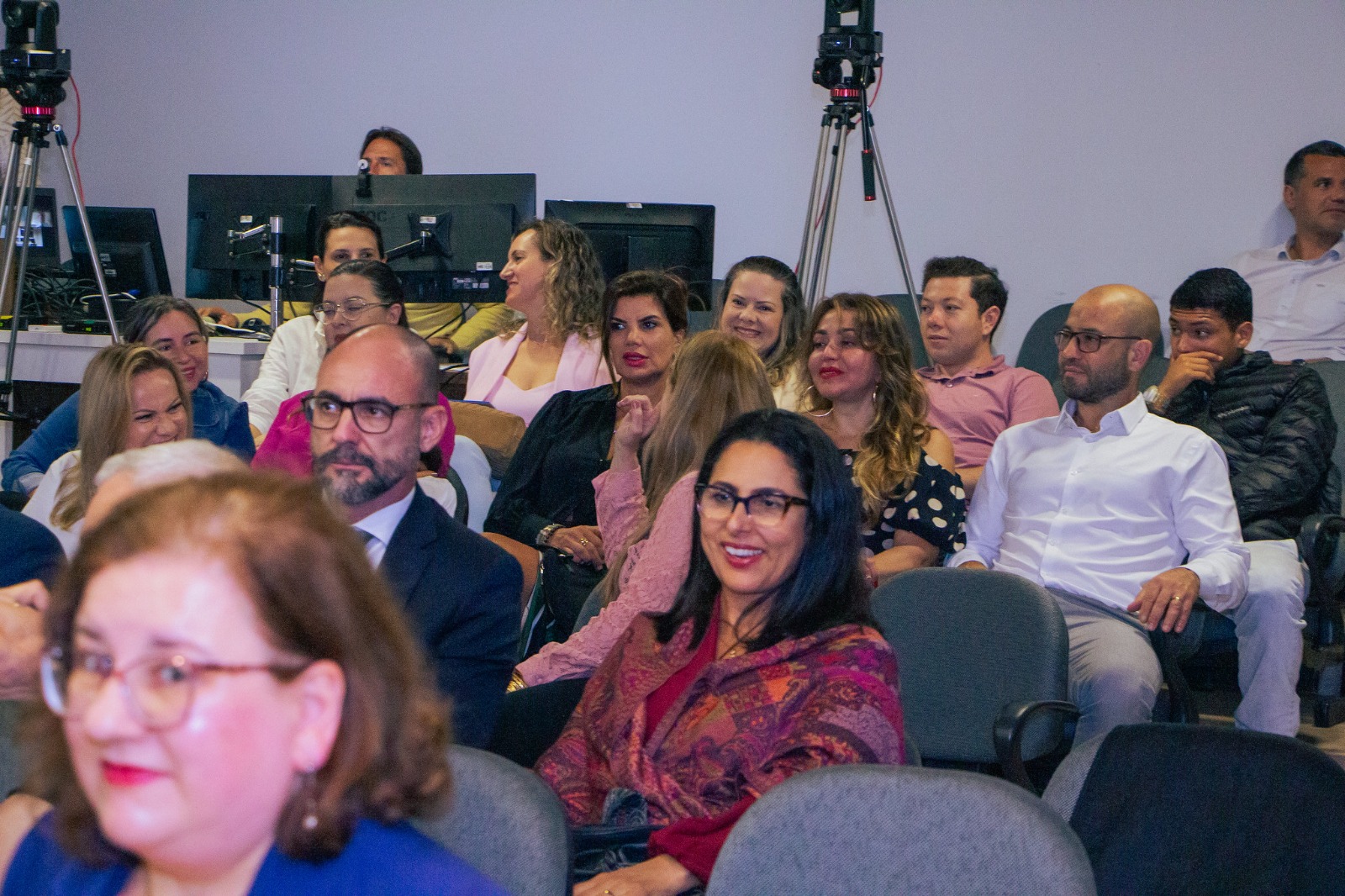 pessoas sorridentes, vestidas formalmente, sentadas em cadeiras de auditório