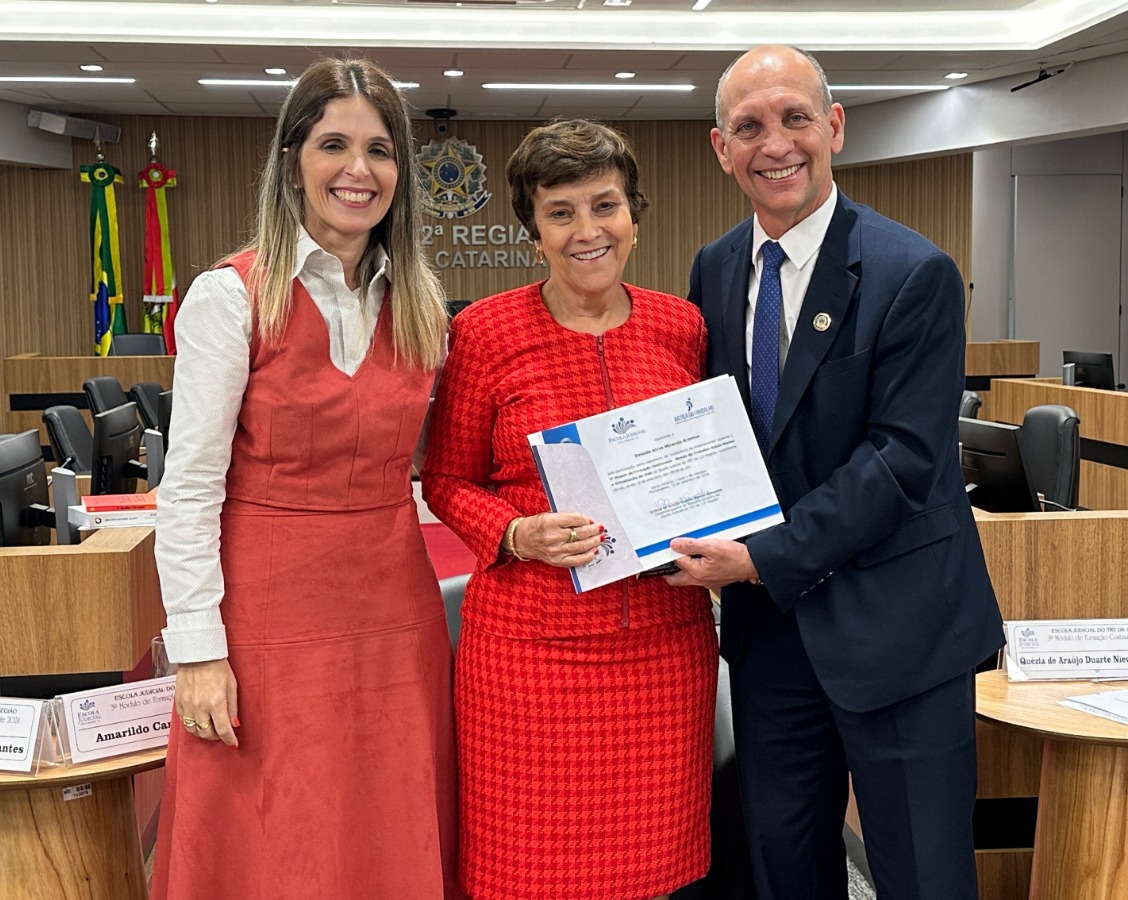 Um homem e duas mulheres olham para a câmera e sorriem. Eles estão vestidos formalmente. O homem segura um certificado e entrega a uma das mulheres.