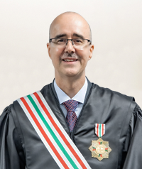 Uma homem de careca e de óculos, vestindo uma toga e uma faixa está sorrindo para a foto