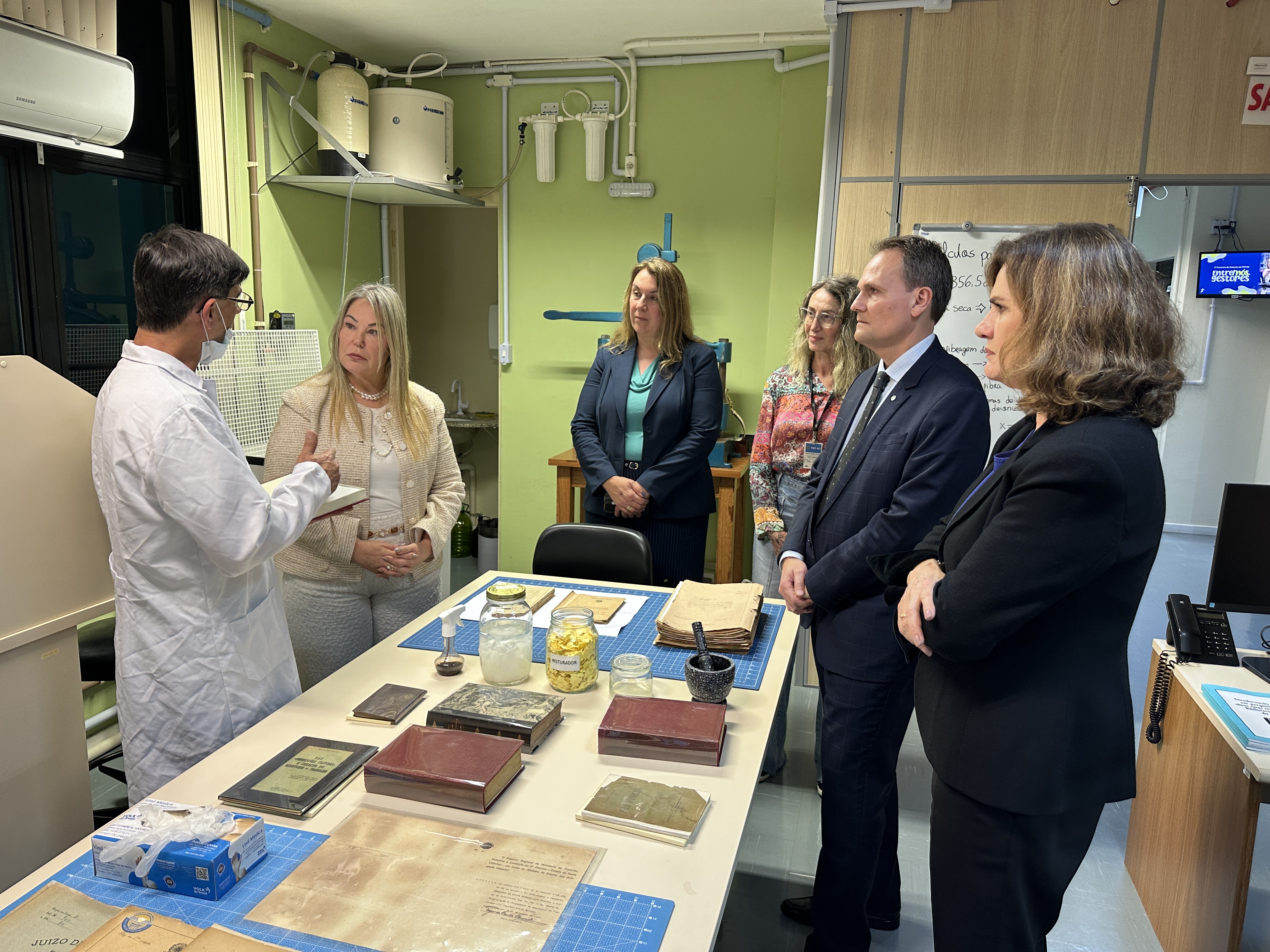 Quatro mulheres e dois homens estão em pé, ao redor de uma mesa de laboratório de documentos