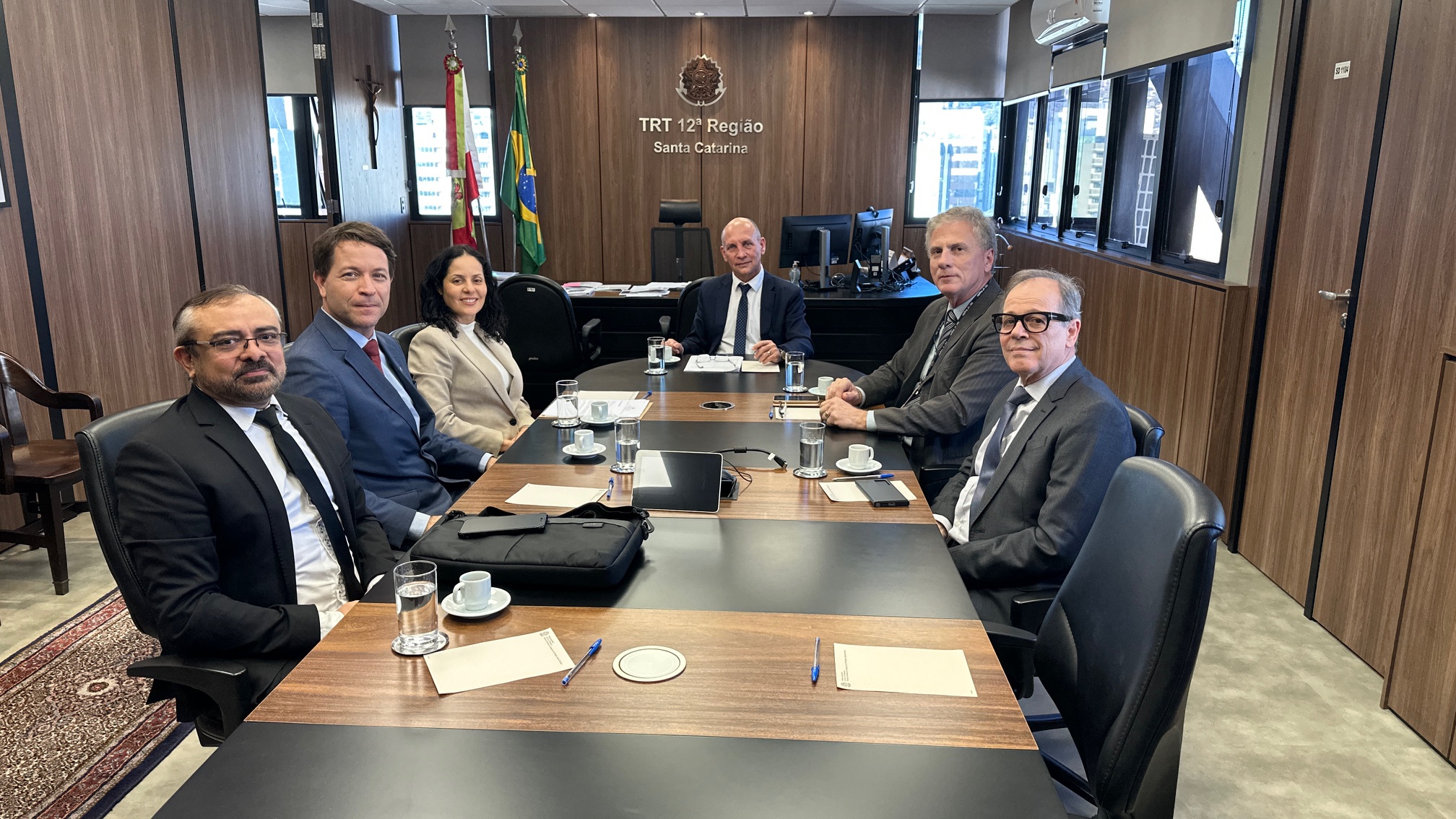 Cinco homens e uma mulher estão sentados em uma mesa de reunião oval, posando para a foto
