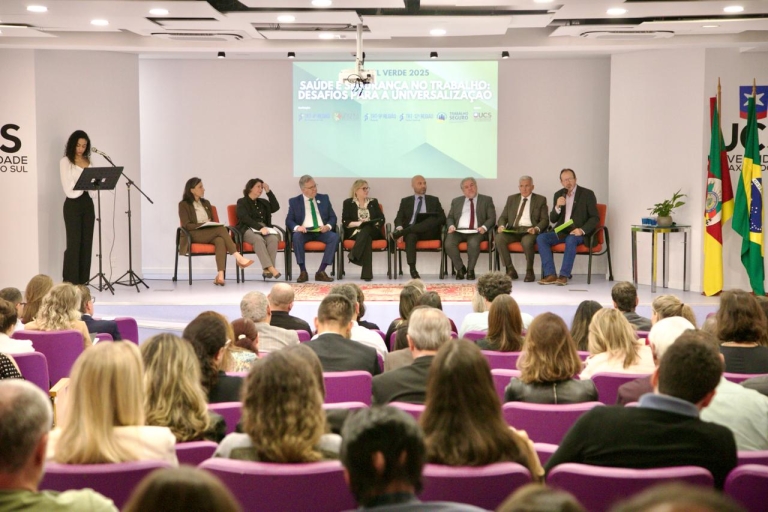 Oito pessoas sentadas em cadeiras, sobre um palco, diante de uma plateia