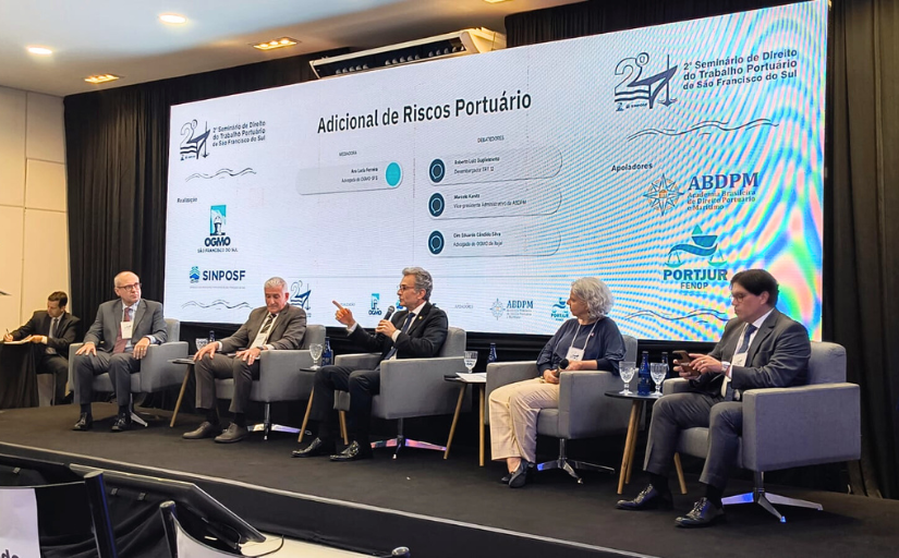 A imagem mostra um painel de discussão em um evento formal. Cinco pessoas estão sentadas no palco, com microfones à frente, participando do evento. 