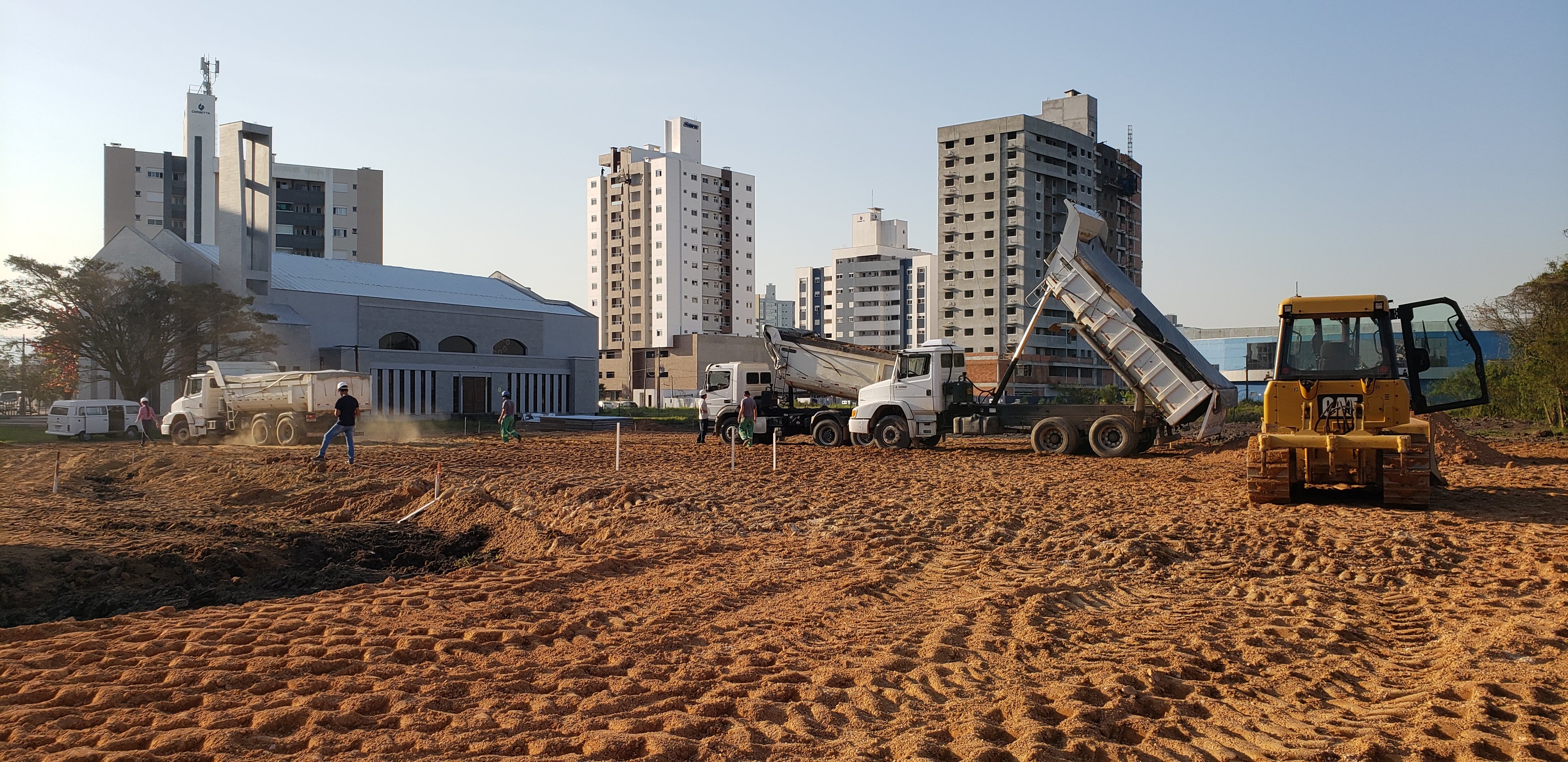 Imagem geral do terreno com caminhões basculantes e trator de esteiras trabalhando na terraplenagem