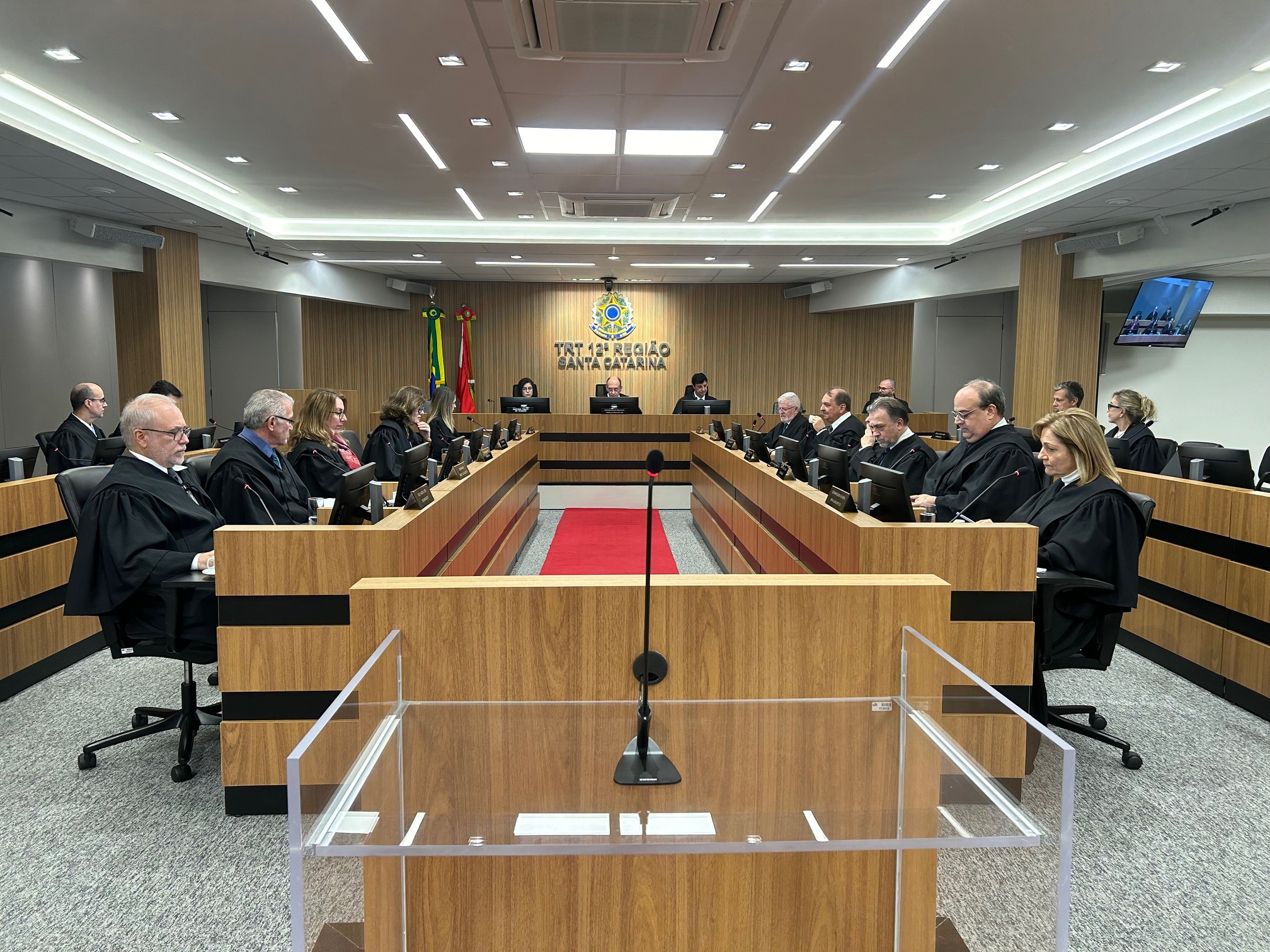 Foto de uma sala de sessões de julgamento, com 18 juízes e juízas dispostos em duas filas laterais e uma ao fundo.
