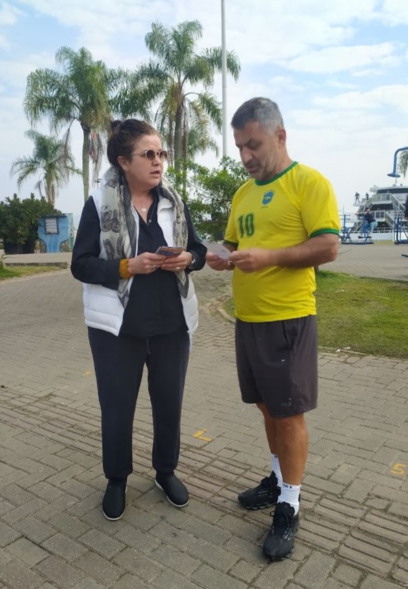 Foto de uma mulher branca de cabelo escuro amarrado em coque, vestindo colete branco camisa preta e óculos escuros, e um homem branco de cabelo curto grisalho, vestido em roupas esportivas e uma camisa verde e amarela da seleção brasileira. Ele olha para um panfleto que tem nas mãos e que a mulher também carrega. O ambiente é aberto