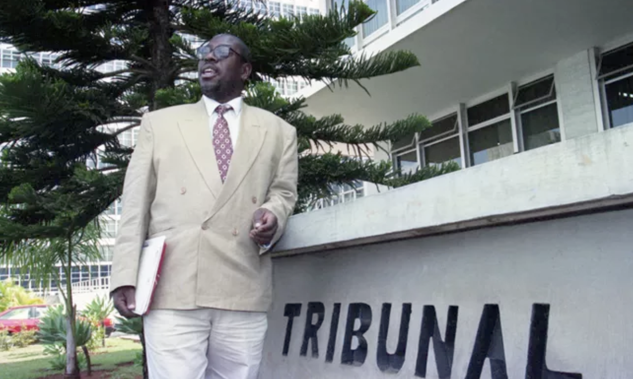 Homem negro de terno claro e gravata estampada está em pé diante de um prédio público com a palavra “TRIBUNAL” na fachada