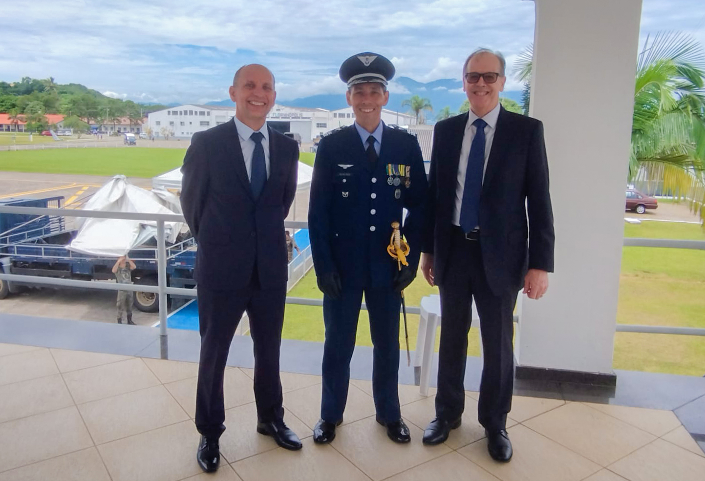 Três homens de pé, dois de terno e gravata e um de farda azul da aeronáutica, posam para a foto