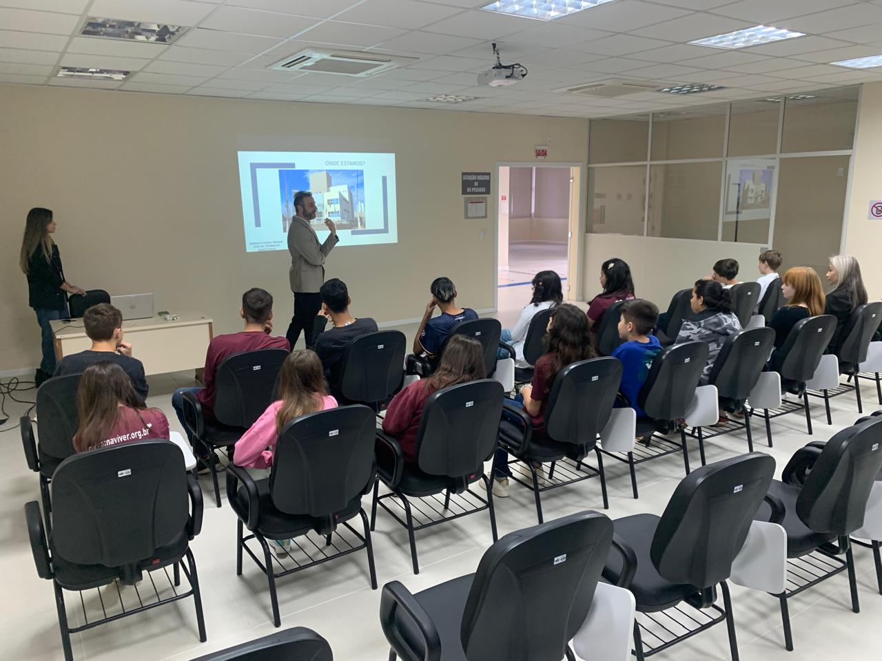 Jovens sentados assistindo um homem em pé, de paletó bege, dando uma palestra