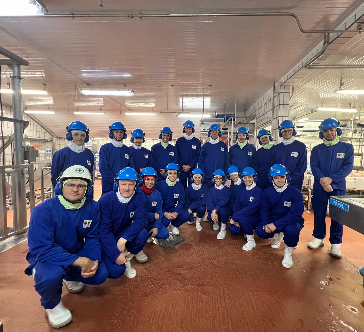 Grupo de pessoas vestidas de macacões azuis, capacetes e fones de ouvido dentro de uma fábrica