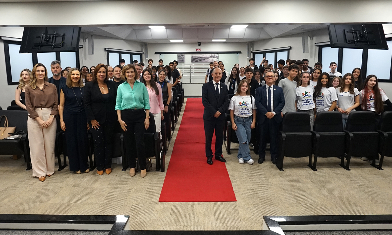 Cerca de 100 pessoas em pé reunidas em um auditório em uma sala ampla sala de julgamento