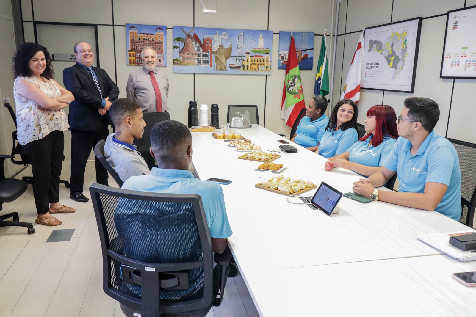 Uma sala de reunião com dois homens e uma mulher em pé, mais três homens e três mulheres sentadas. No meio da mesa tem salgadinhos e um tablet.