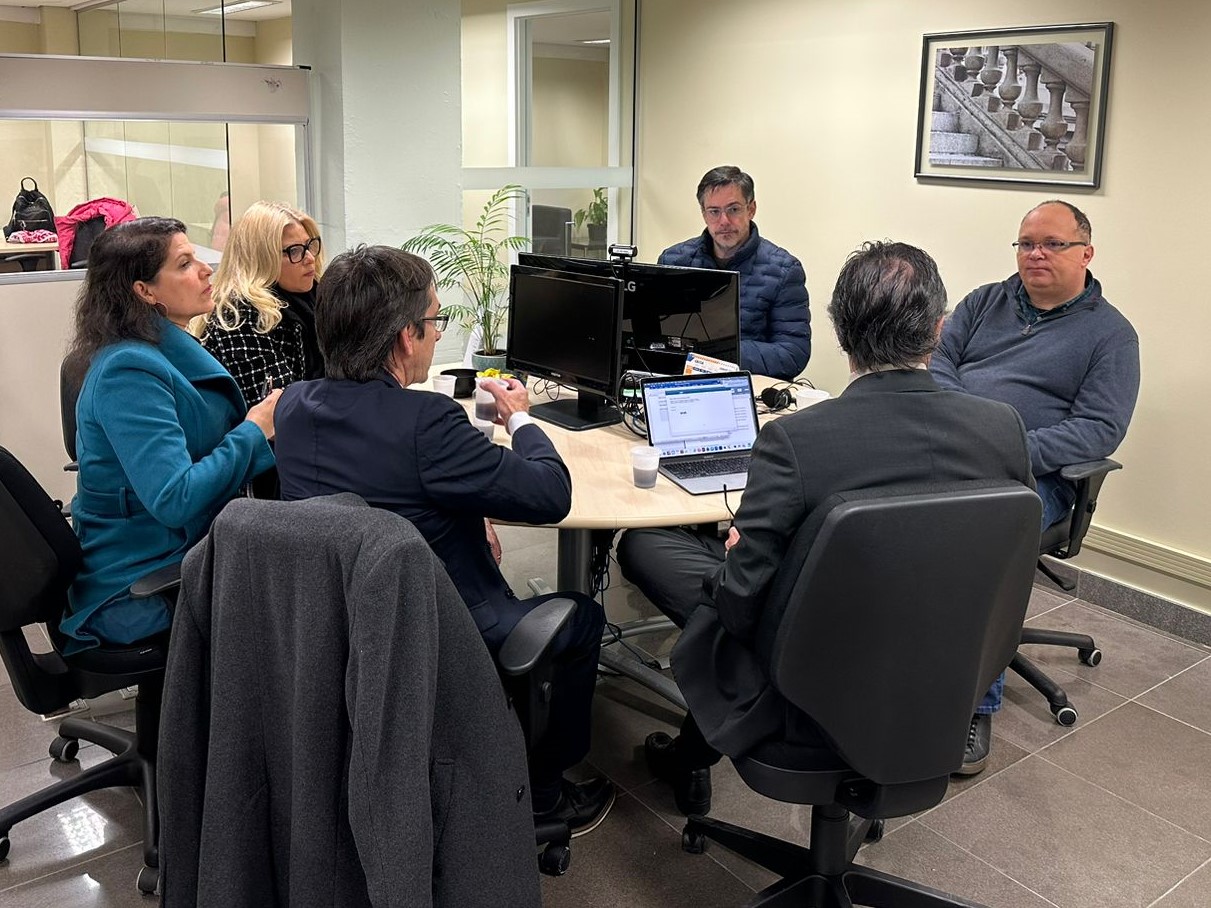 Foto mostra seis pessoas ao redor de uma mesa redonda. Eles parecem estar conversando e debatendo.