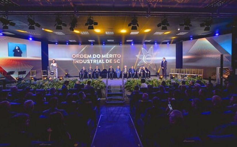 Cerimônia formal com autoridades no palco e plateia ocupada; ao fundo, projeção com o texto “Ordem do Mérito Industrial de SC” e intérprete de Libras visível à esquerda.