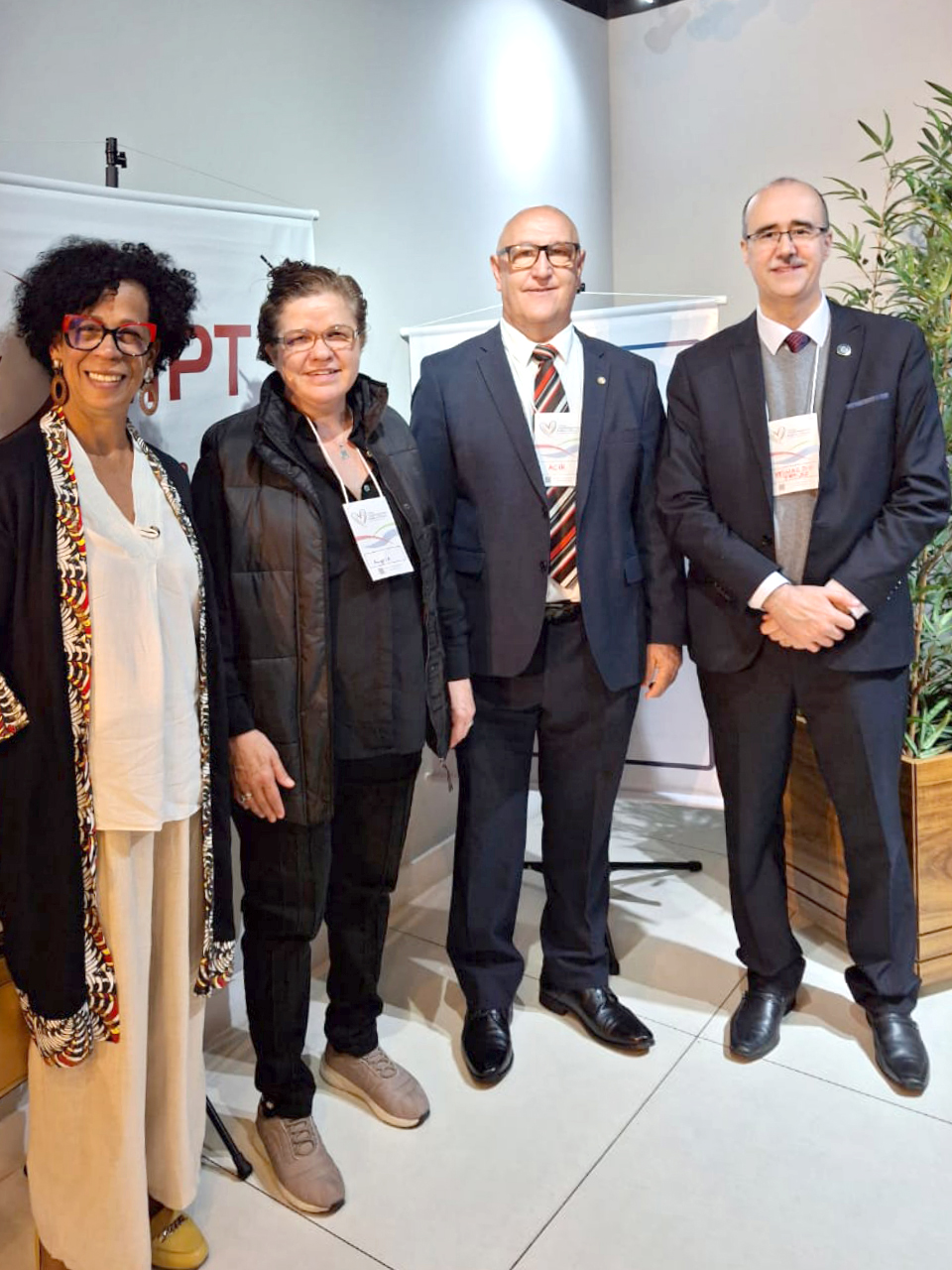 A imagem retrata quatro pessoas posando para uma foto em um ambiente formal, provavelmente relacionado a um evento institucional. Duas mulheres e dois homens estão lado a lado, todos usando crachás de identificação. As mulheres estão à esquerda, uma delas vestida com um casaco colorido e a outra com uma jaqueta preta. Os dois homens à direita usam ternos escuros e gravatas. Todos sorriem para a câmera, e ao fundo há um banner que parece conter siglas de uma organização ou evento. O ambiente tem plantas decorativas e uma iluminação clara.