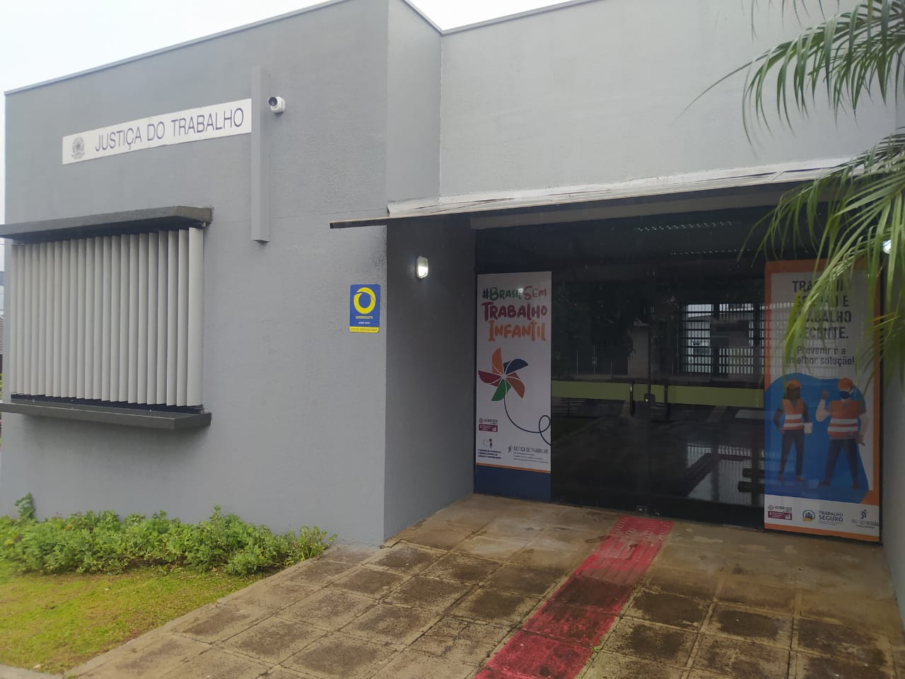 Entrada do prédio da Vara do Trabalho de Xanxerê, com fachada em concreto, banners contra o trabalho infantil, piso de concreto e uma pequena área verde ao lado.