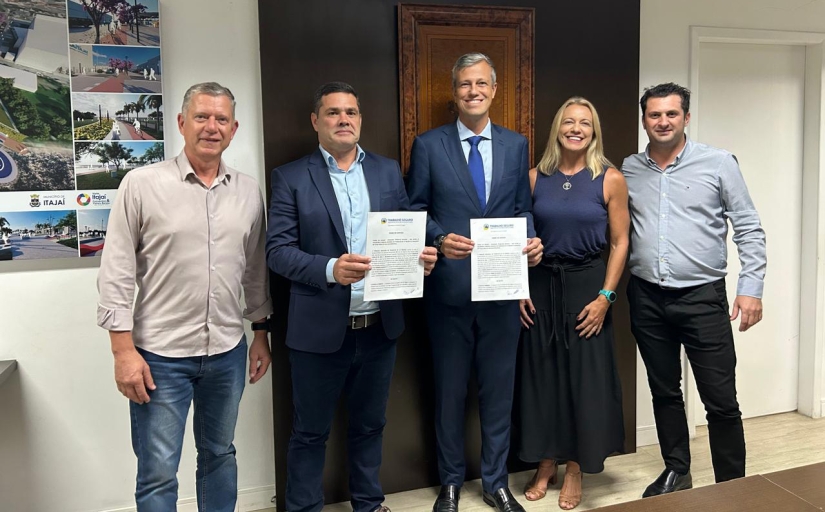Grupo de cinco pessoas posam sorridentes, em pé, para a foto. Dois deles, ao centro, mostram um documento
