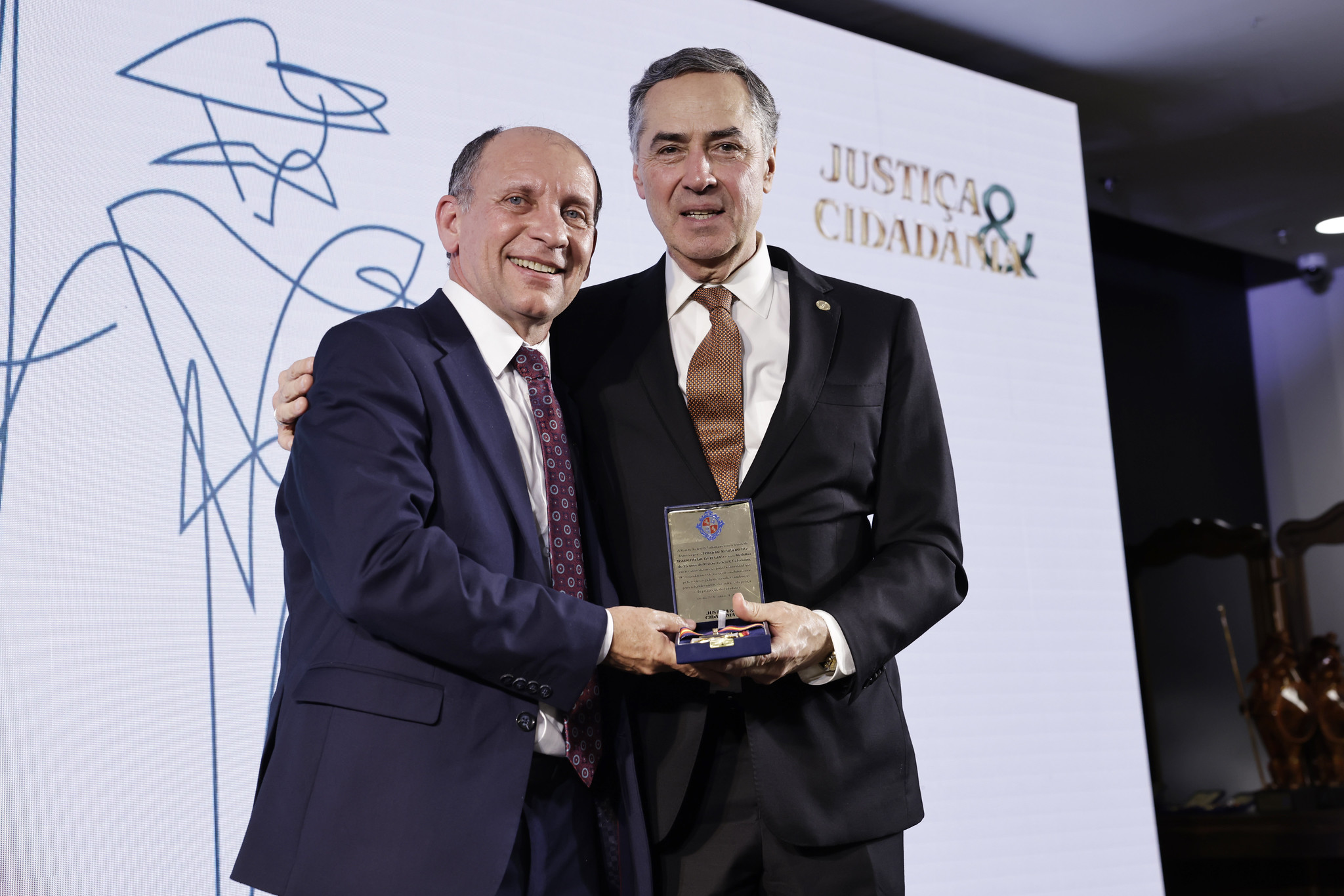 Dois homens brancos, em trajes formais, em cima de um palco, sorriem para a foto. Um deles entrega ao outro uma medalha em mãos.