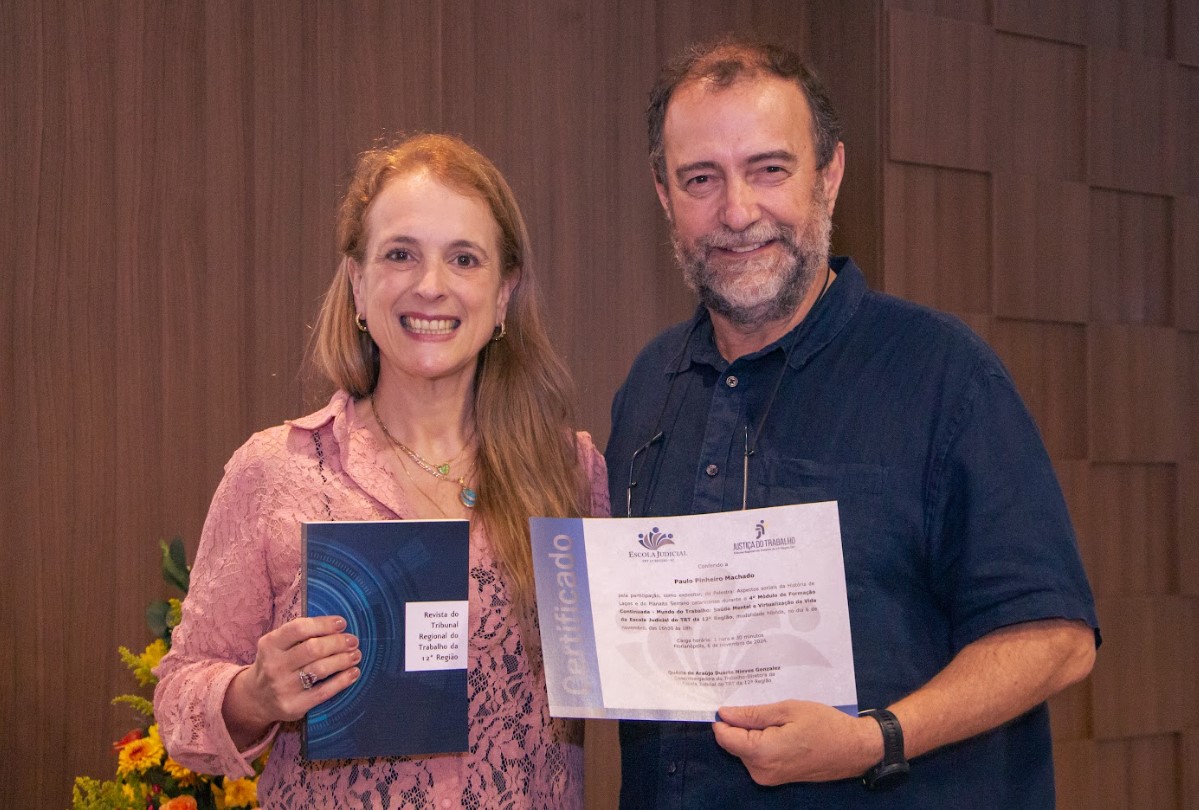 Um homem e uma mulher posam para a foto. Ela segura um livro e ele um certificado