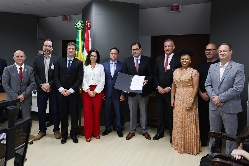 Um grupo de dez pessoas, todas vestidas formalmente, posa em um ambiente formal. O homem no centro segura um documento aberto. Bandeiras estão ao fundo, sugerindo um evento oficial.