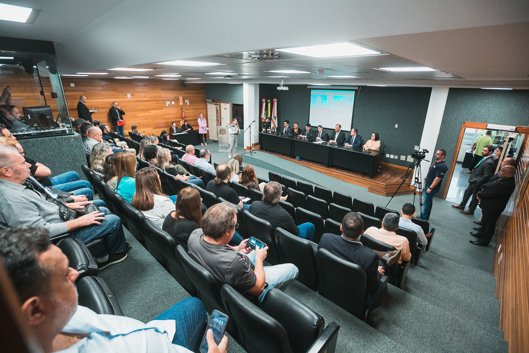 Foto aberta mostrando um ambiente de evento. Seis pessoas sentadas na mesa principal sendo assistidas por um grupo de cerca de outras 40 pessoas em um auditório.