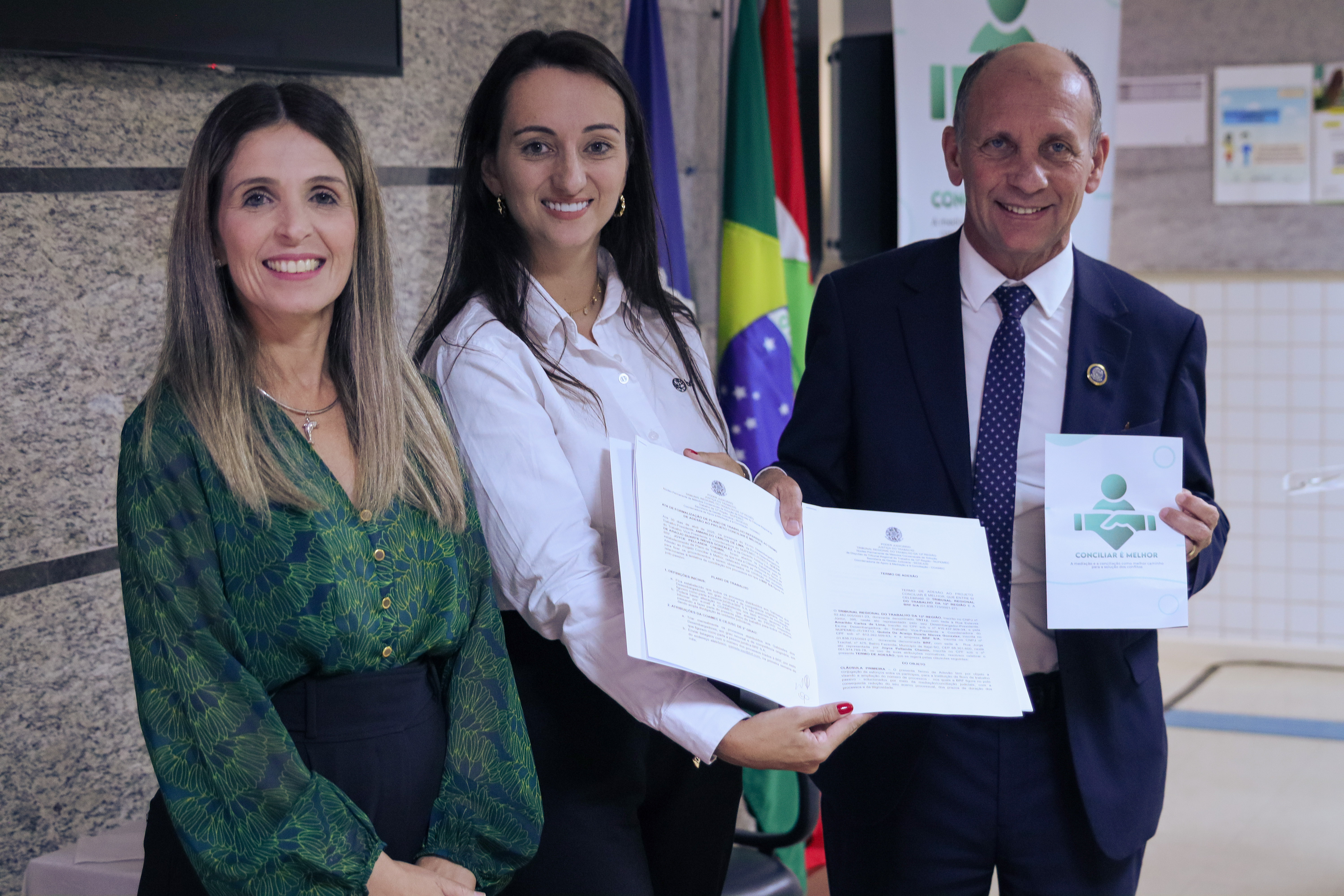 Três pessoas em pé, sendo duas mulheres e um homem, sorriem para a foto. Uma das mulheres, ao centro, de pele branca e cabelos longos e pretos, trajando uma camisa branca e calça preta, segura nas mãos uma pasta com um documento assinado.