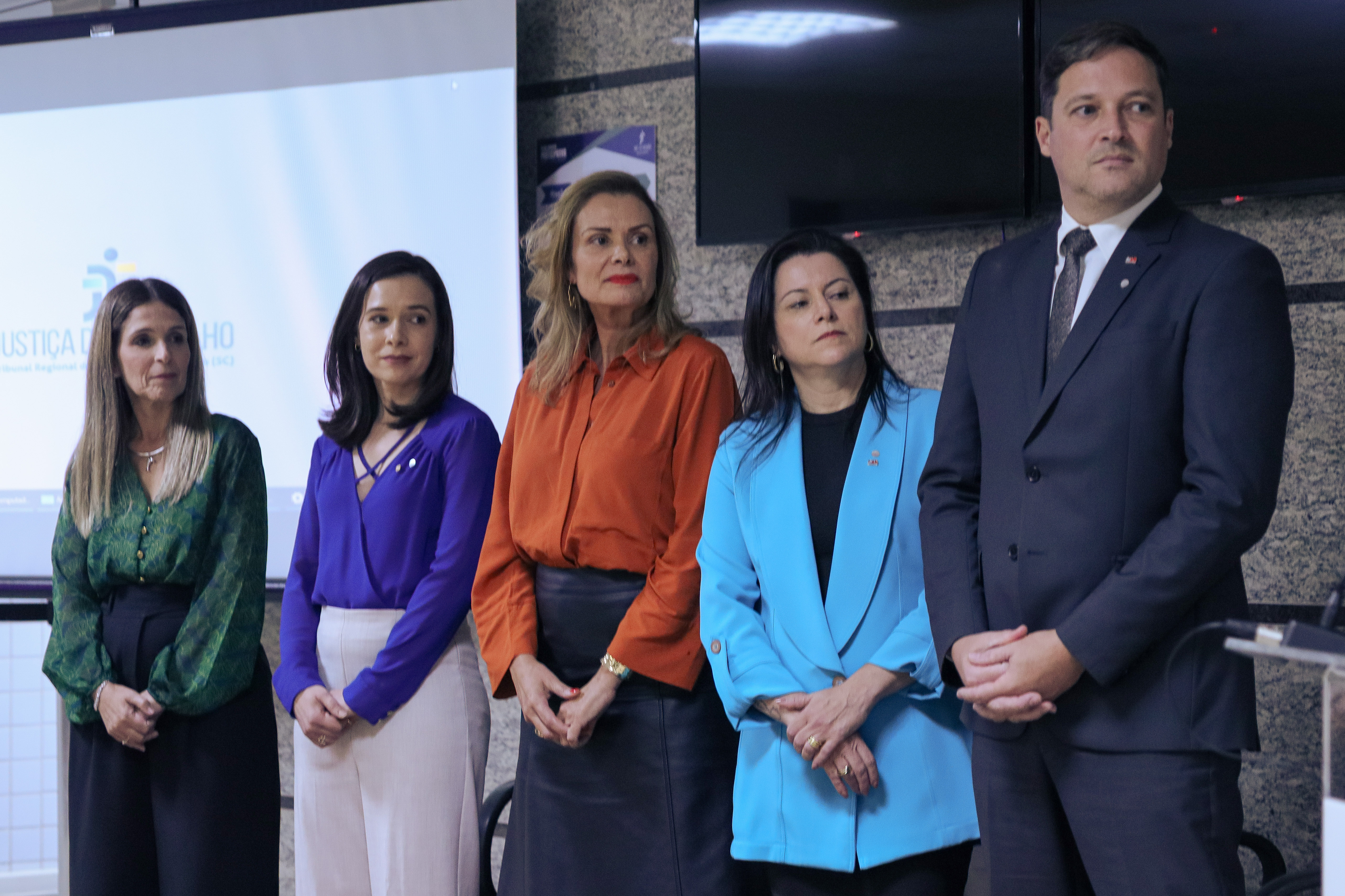 Cinco pessoas, em pé, em trajes formais, estão em um palco de auditório. São quatro mulheres e um homem.