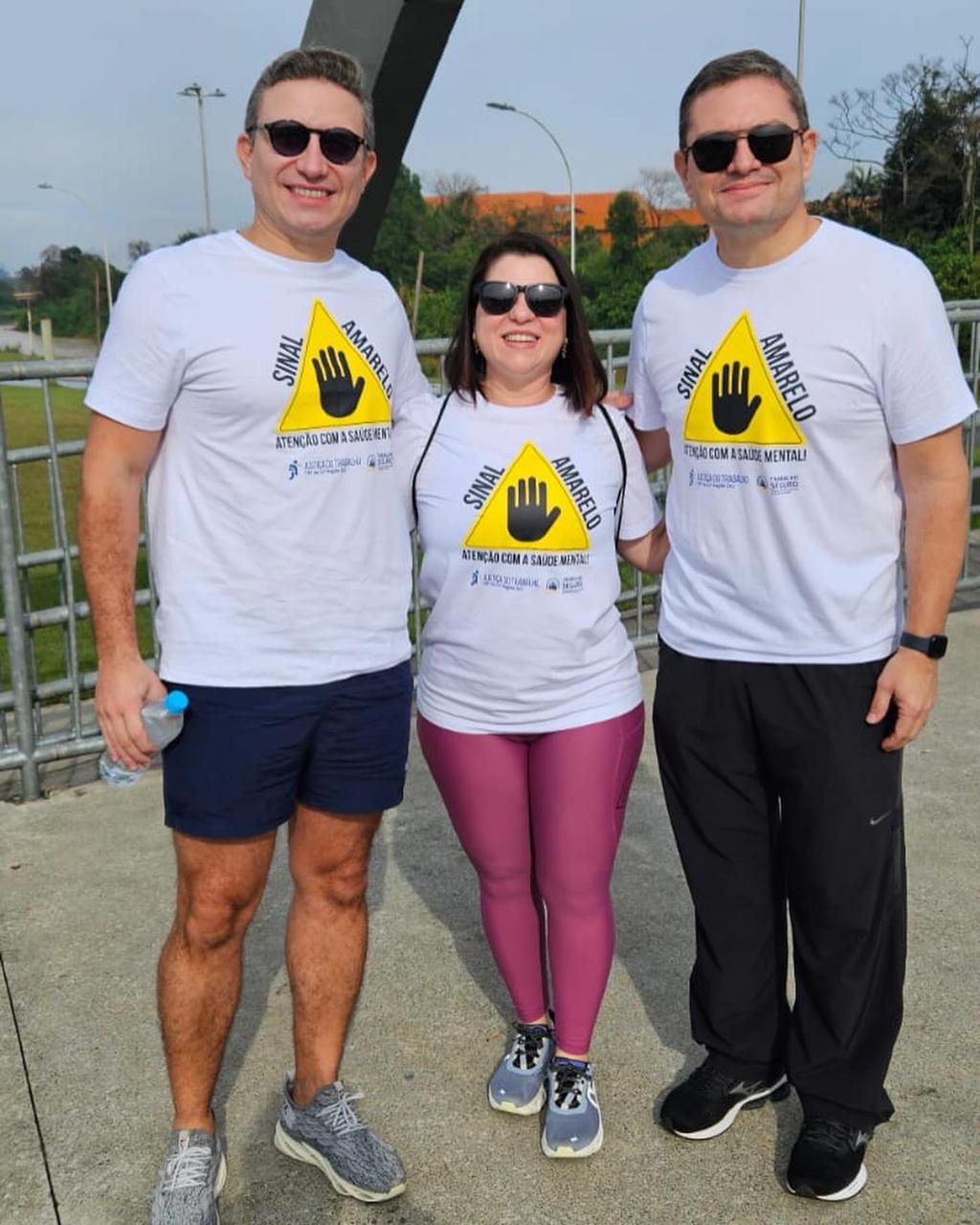 A imagem mostra três pessoas posando para uma foto em uma área ao ar livre. Todos estão usando camisetas brancas com o símbolo "Sinal Amarelo" e a frase "Atenção com a Saúde Mental"