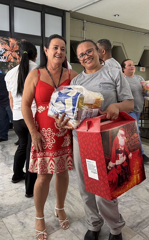 Duas mulheres sorriem, em pé, para a foto. Uma delas segura uma cesta de Natal e uma cesta básica de alimentos.