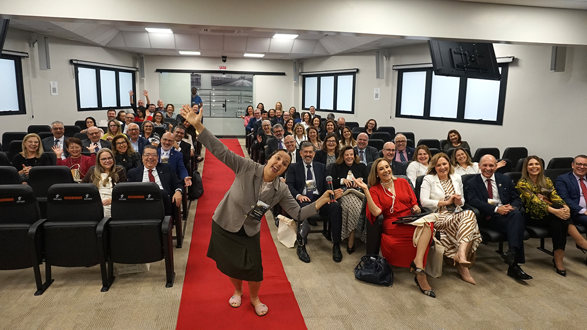 Grupo de pessoas em auditório sorrindo para foto, com mulher à frente animada segurando microfone.