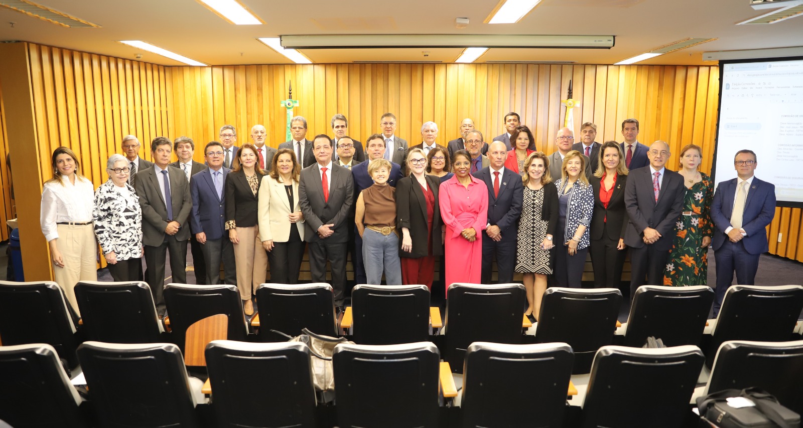 35 pessoas em pé, lado a lado, posam para a foto. Os homens vestem terno, e as mulheres, vestuário diversificado. Ao fundo, um painel de MDF dourado e iluminado