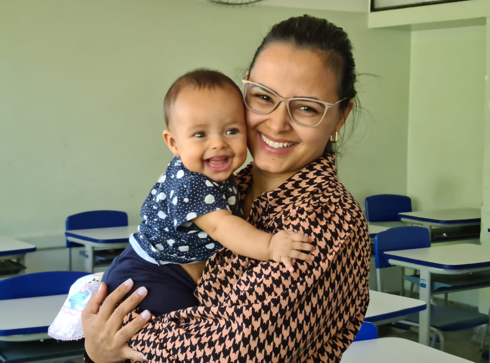 Foto mostra mãe sorridente, de óculos, segurando um bebê sorridente no colo