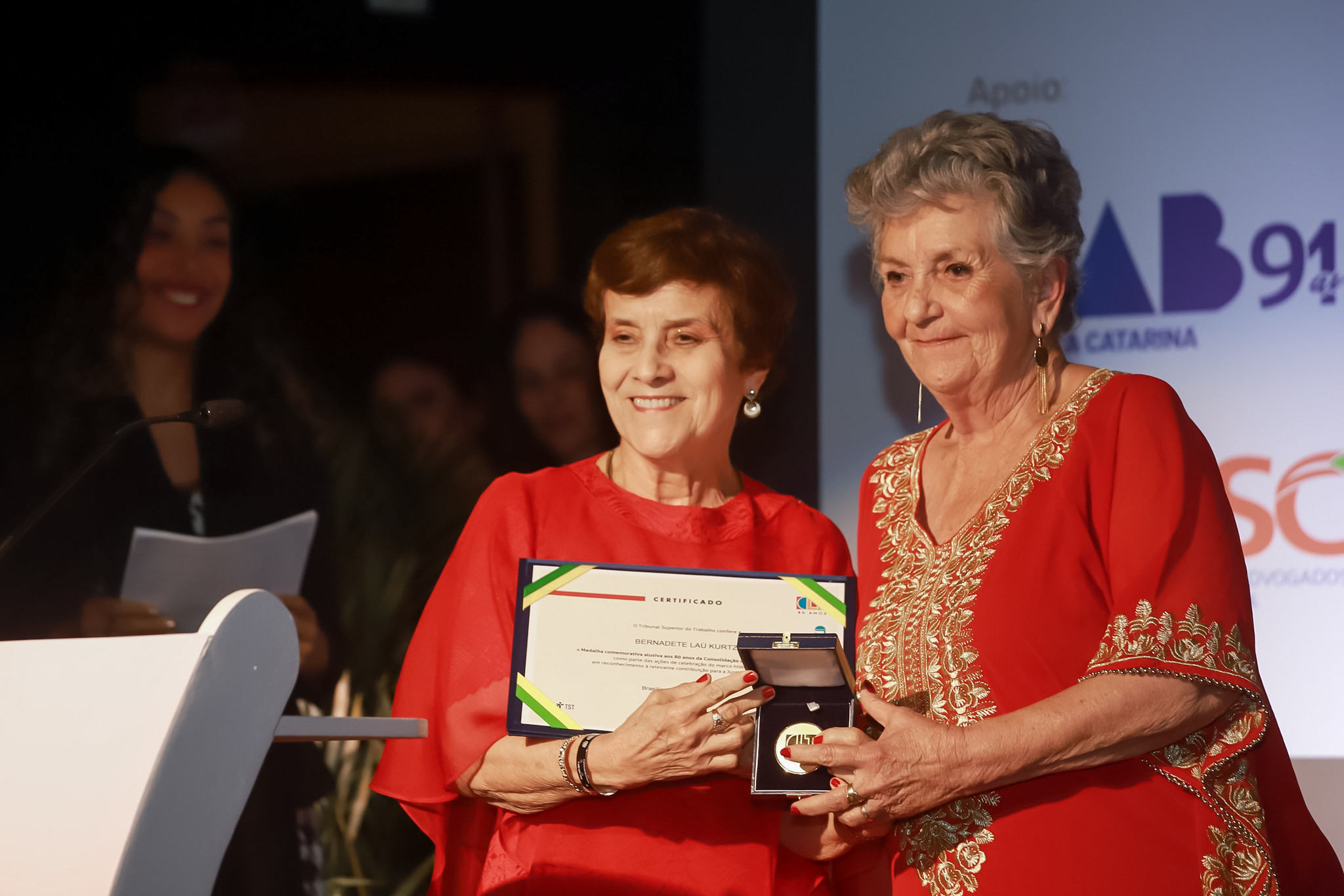 Duas mulheres idosas, de cabelos curtos, ambas trajando roupas vermelhas, posam para a foto. Uma delas, à esquerda, segura um certificado enquanto a que está à direita entrega uma medalha.