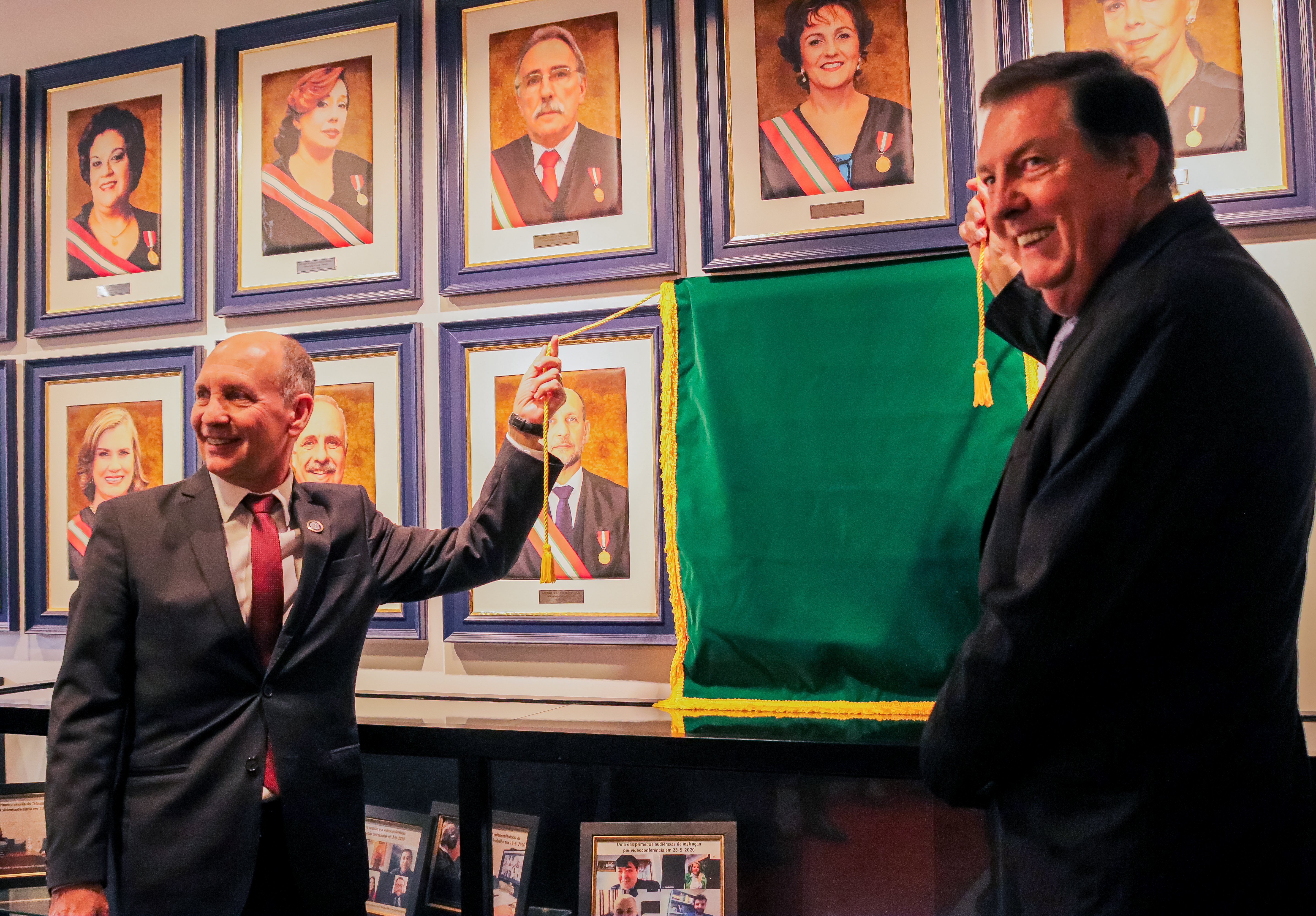 Dois homens sorridentes se preparam para descerrar um quadro, coberto por um pano verde. Ao fundo, diversos quadros de ex-corregedores expostos em uma parede