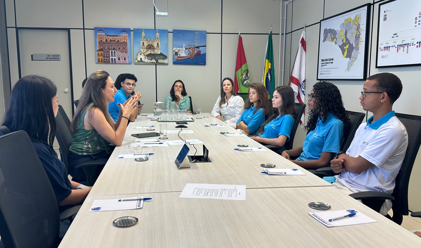 Mesa longa de madeira, com aprendizes, magistradas e a psicóloga ao redor dela.