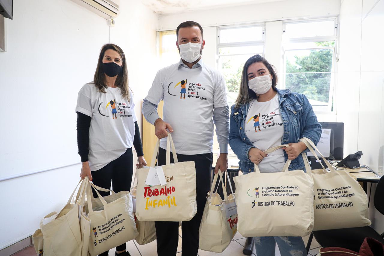 Da esquerda para a direita: servidora Clemair Carneiro, secretário Oscar Silva Neto e servidora da Secretaria lado a lado, em foto posada, com a camiseta do Programa de Combate ao Trabalho Infantil e segurando ecobags cheias com os itens que foram entregues 