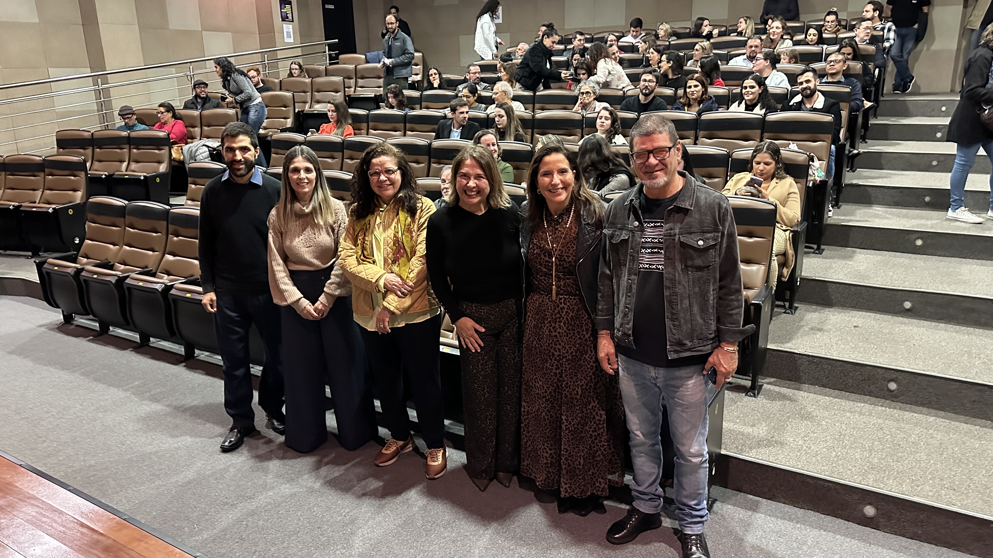 Seies pessoas posam em pé para a foto em um auditório de teatro, sendo dois homens e quatro mulheres. Ao fundo, há pessoas sentadas em cadeiras de auditório.