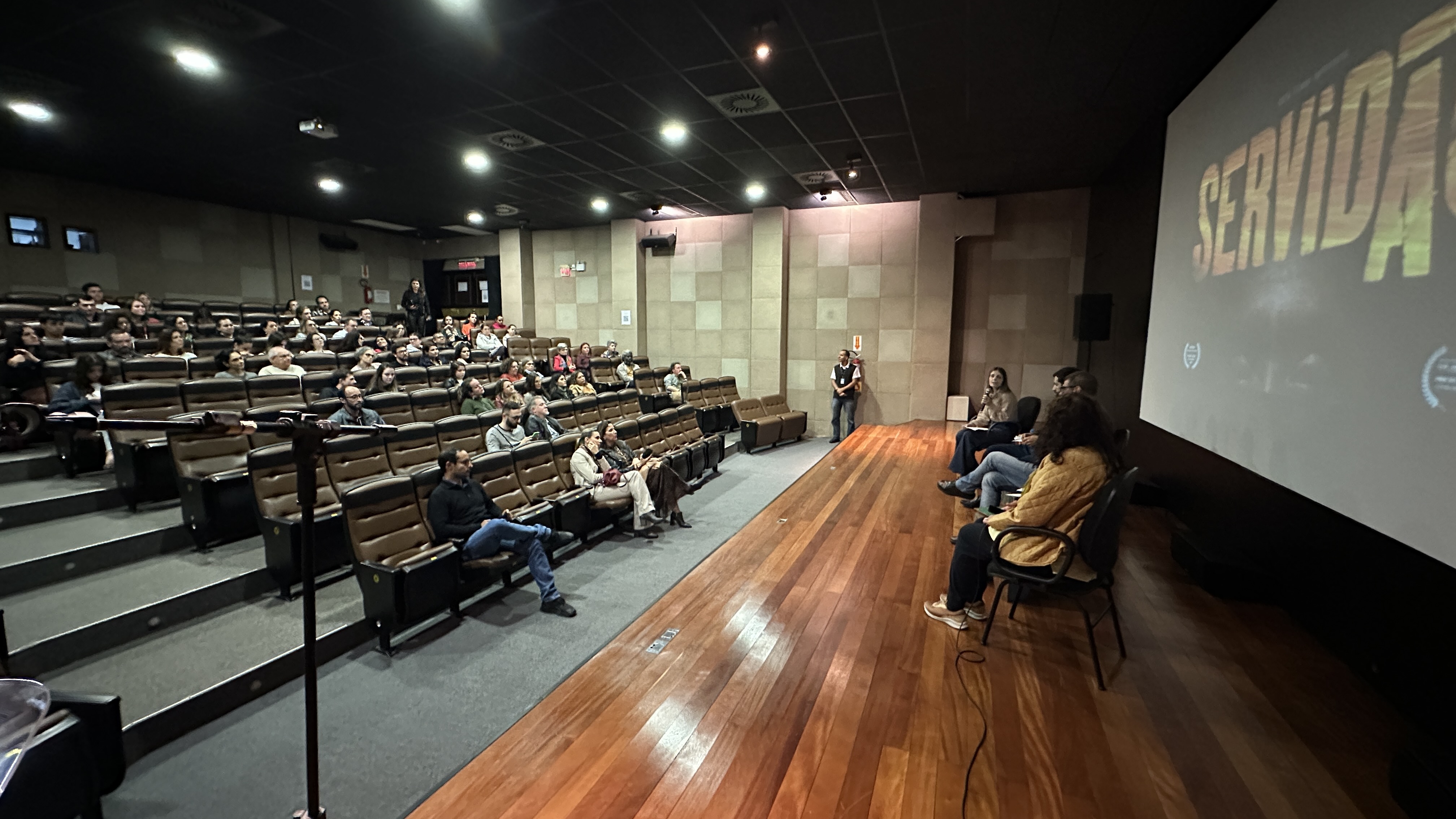 Fotografia de um auditório de teatro. À esquerda, pessoas estão sentadas em cadeiras na plateia. À direita, quatro pessoas estão sentada em cadeiras no palco e, ao fundo, um tela projeta o título do documentário: Servidão