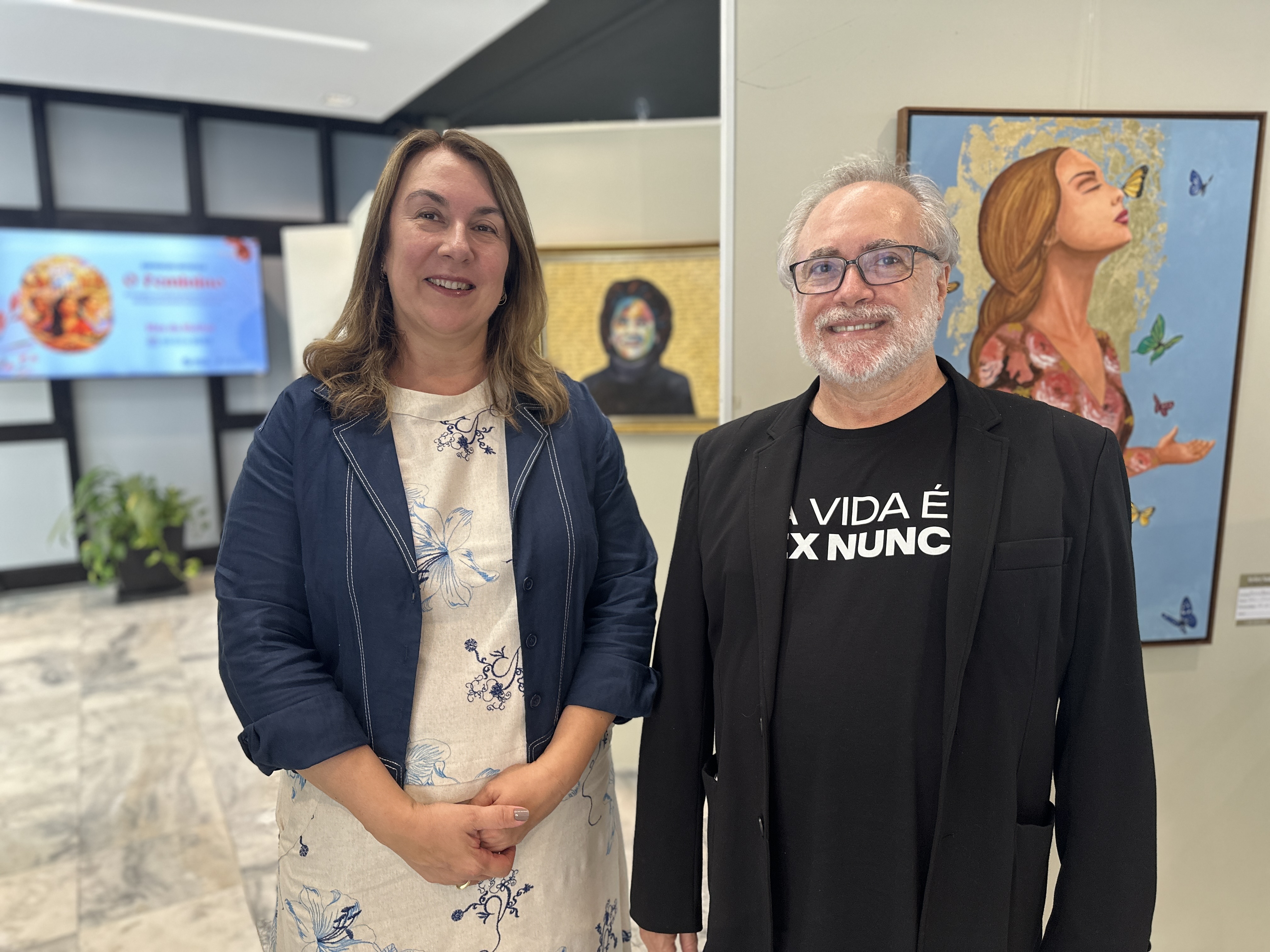 Uma mulher vestida de forma descontraída e um homem vestindo camisa e blazer pretos lado a lado, de pé. Ao fundo, duas pinturas.