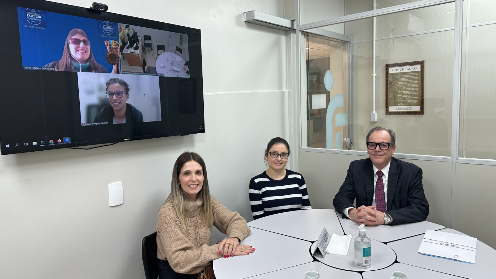 fotografia de três pessoas, duas mulheres e um homem, vestidos formalmente e sentados de frente para uma mesa. ao fundo, um monitor com outras duas mulheres em videoconferência