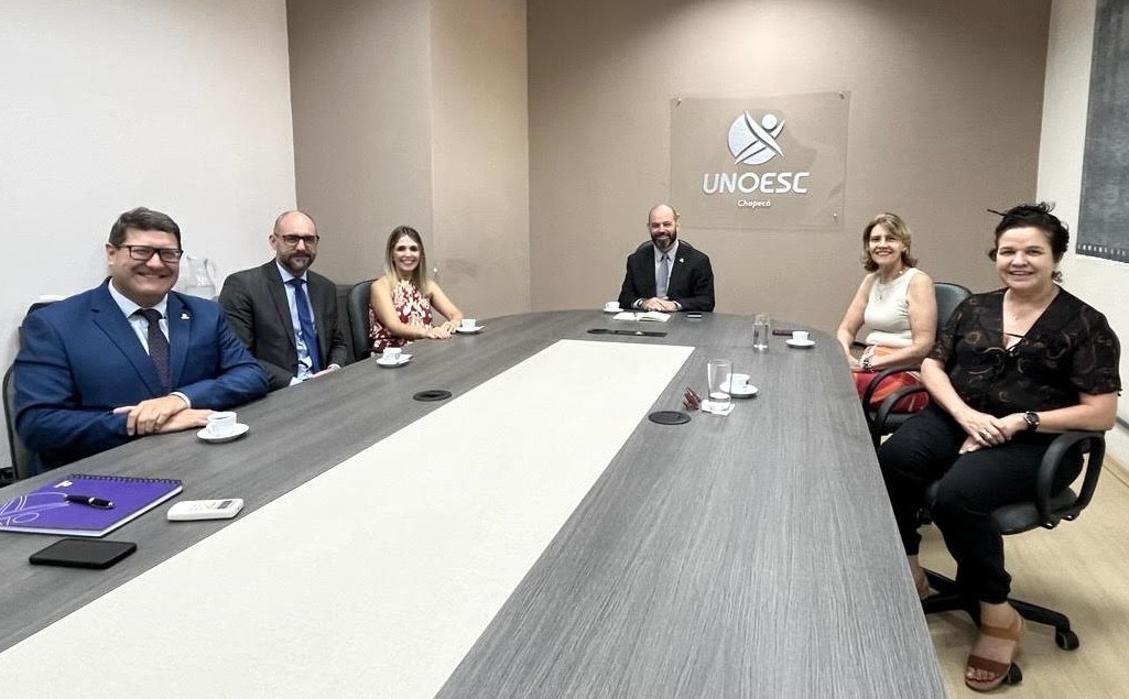 Foto de um grupo de pessoas ao redor de uma mesa de madeira. Ao fundo, a logo da Unoesc