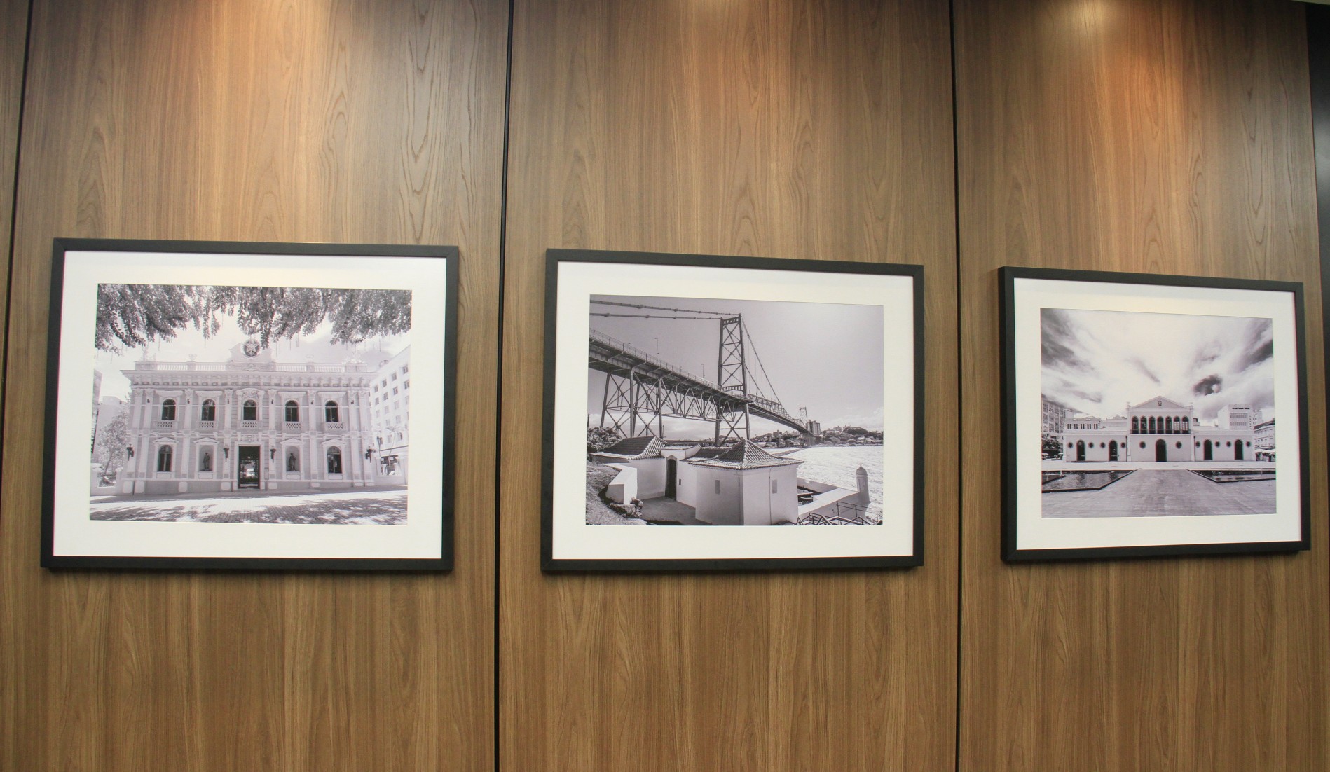 Foto de uma divisória de MDF marrom com três quadros com fotos, em preto e branco, de monumentos históricos de Florianópolis