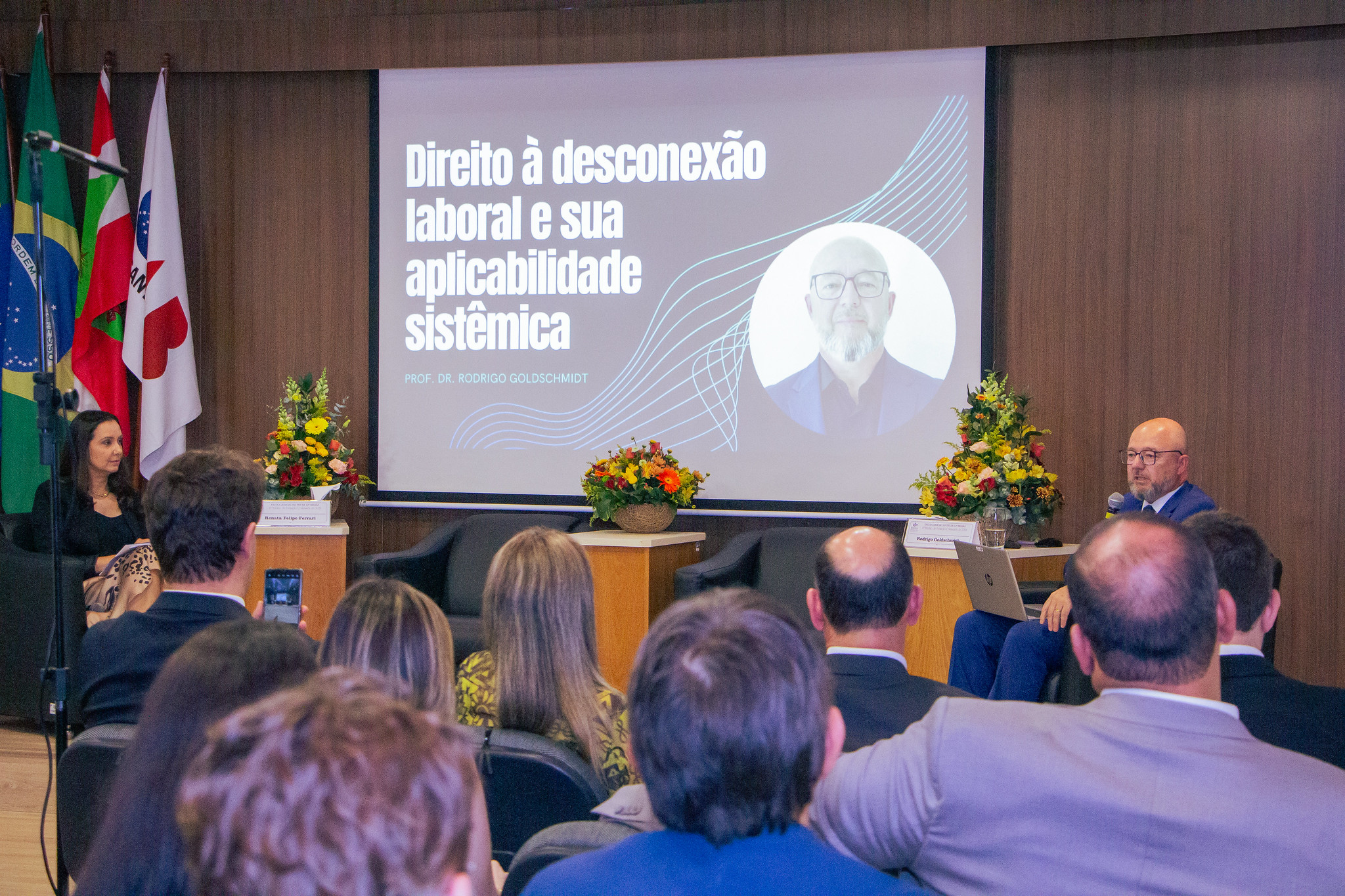 Público de pessoas sentadas, de costas para a foto. Elas estão olhando para duas pessoas à frente, uma delas fala ao microfone